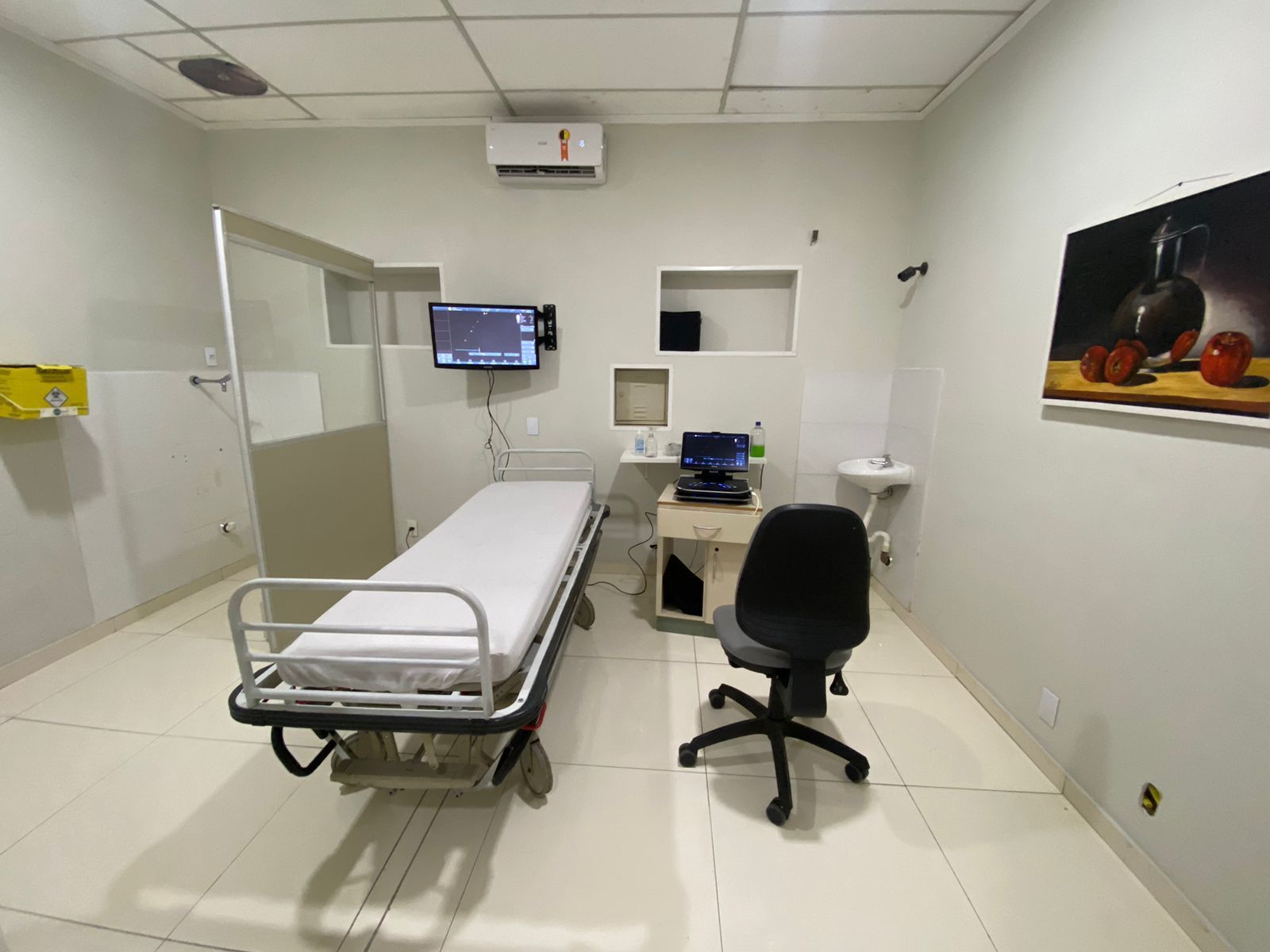 Sala de exame médico: cama, monitor, mesa com equipamentos, obra de arte na parede e cadeira.