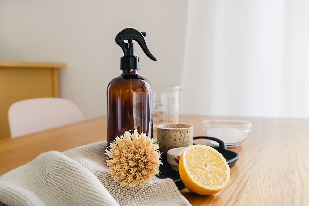 Amber spray bottle, wooden scrub brush, and sliced lemon on a table with towels and glass bowls