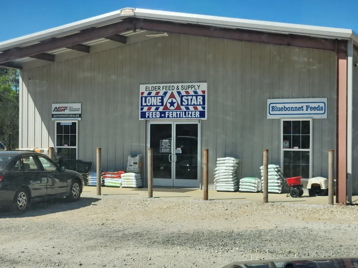 Exterior of a rural feed store with signs, stacks of feed, and a parked truck in front