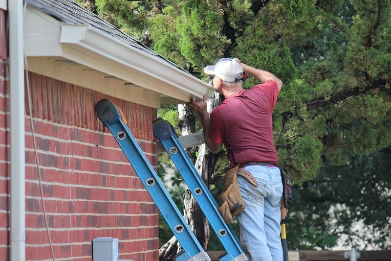 Two workers clean gutters on a house roof using blue ladders.