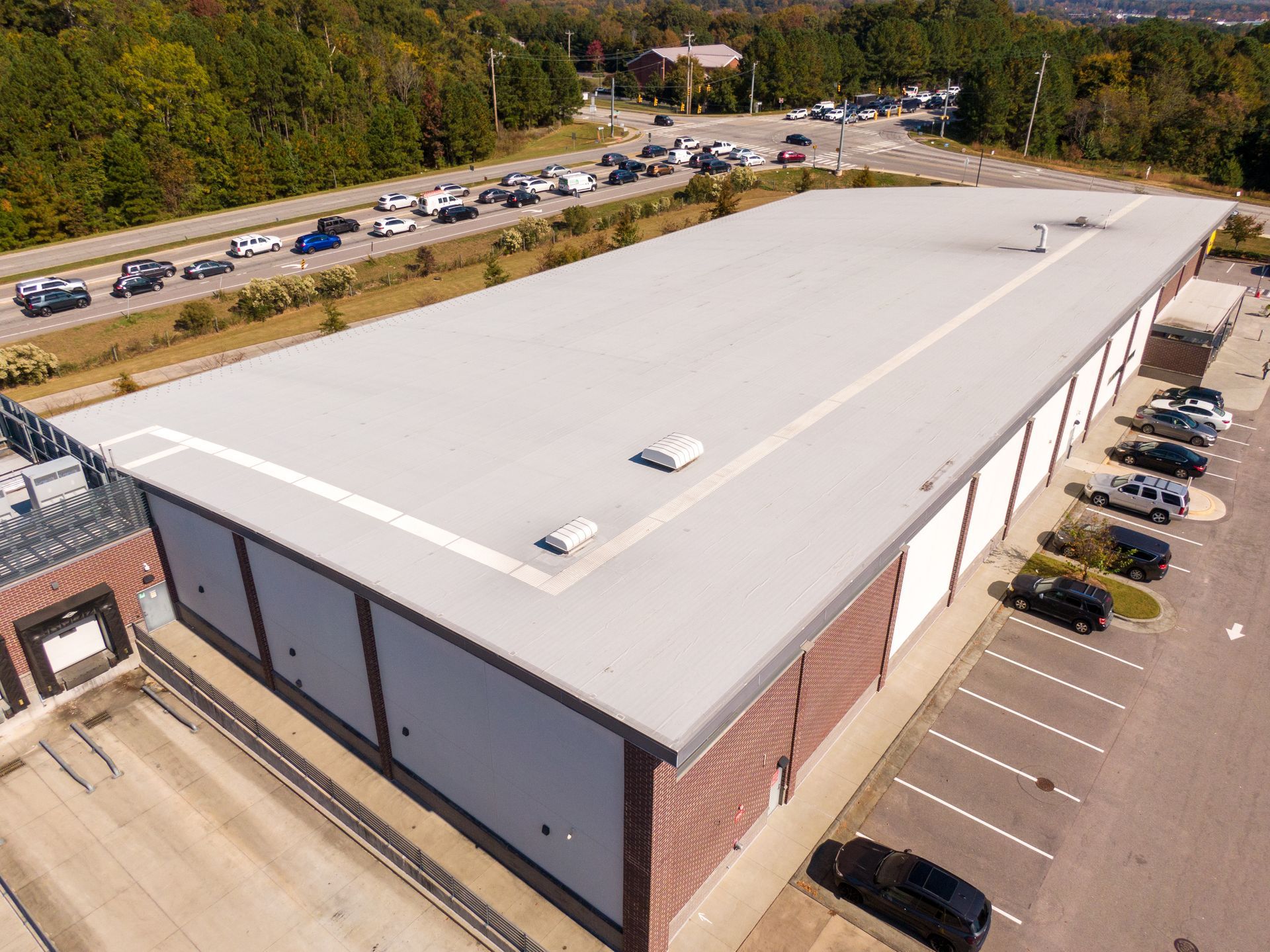 Aerial view of a large white-roofed commercial building with parked cars and trees nearby.