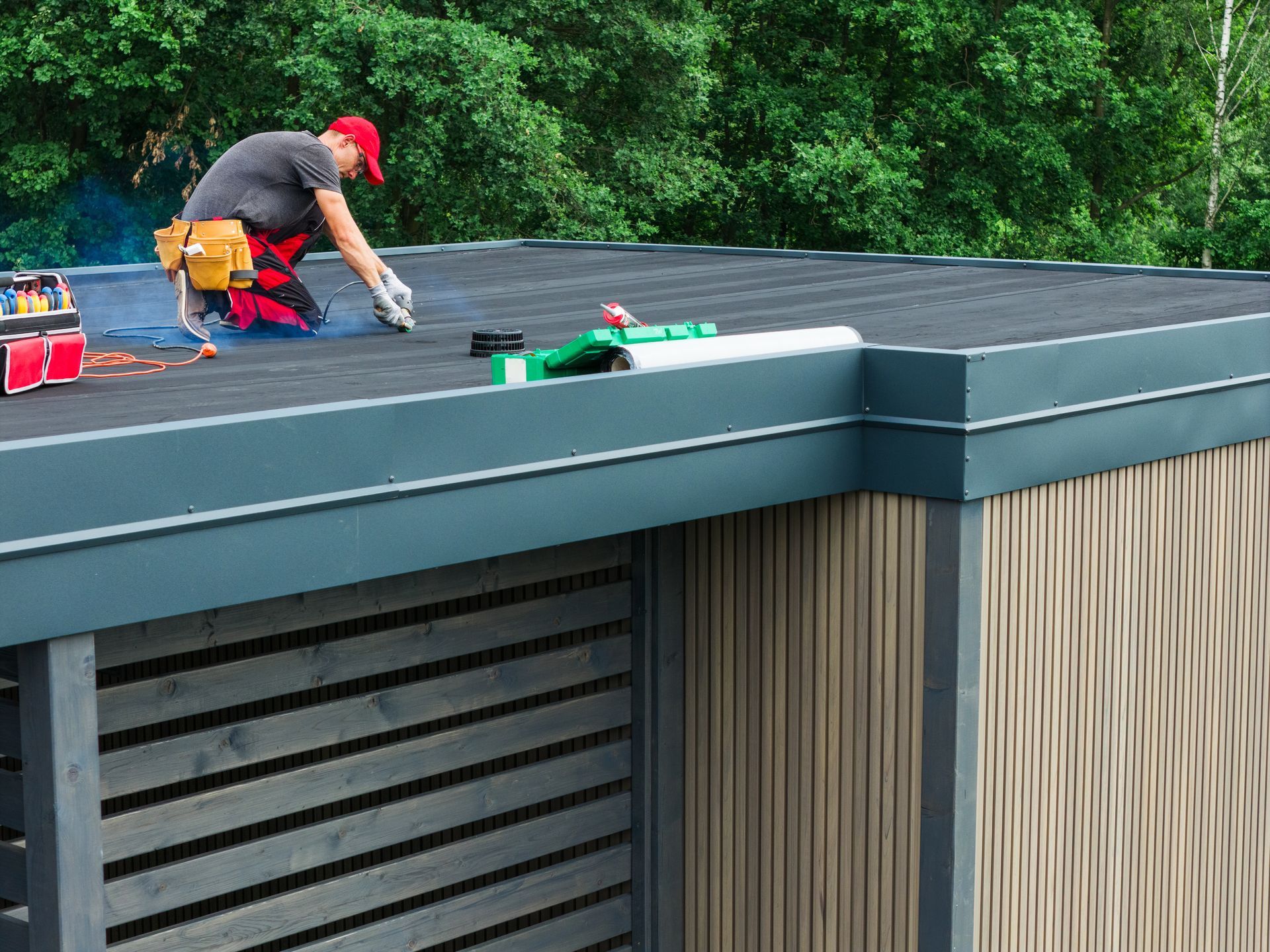 Worker installing a dark roof on a modern building, with tools and green trees in the background