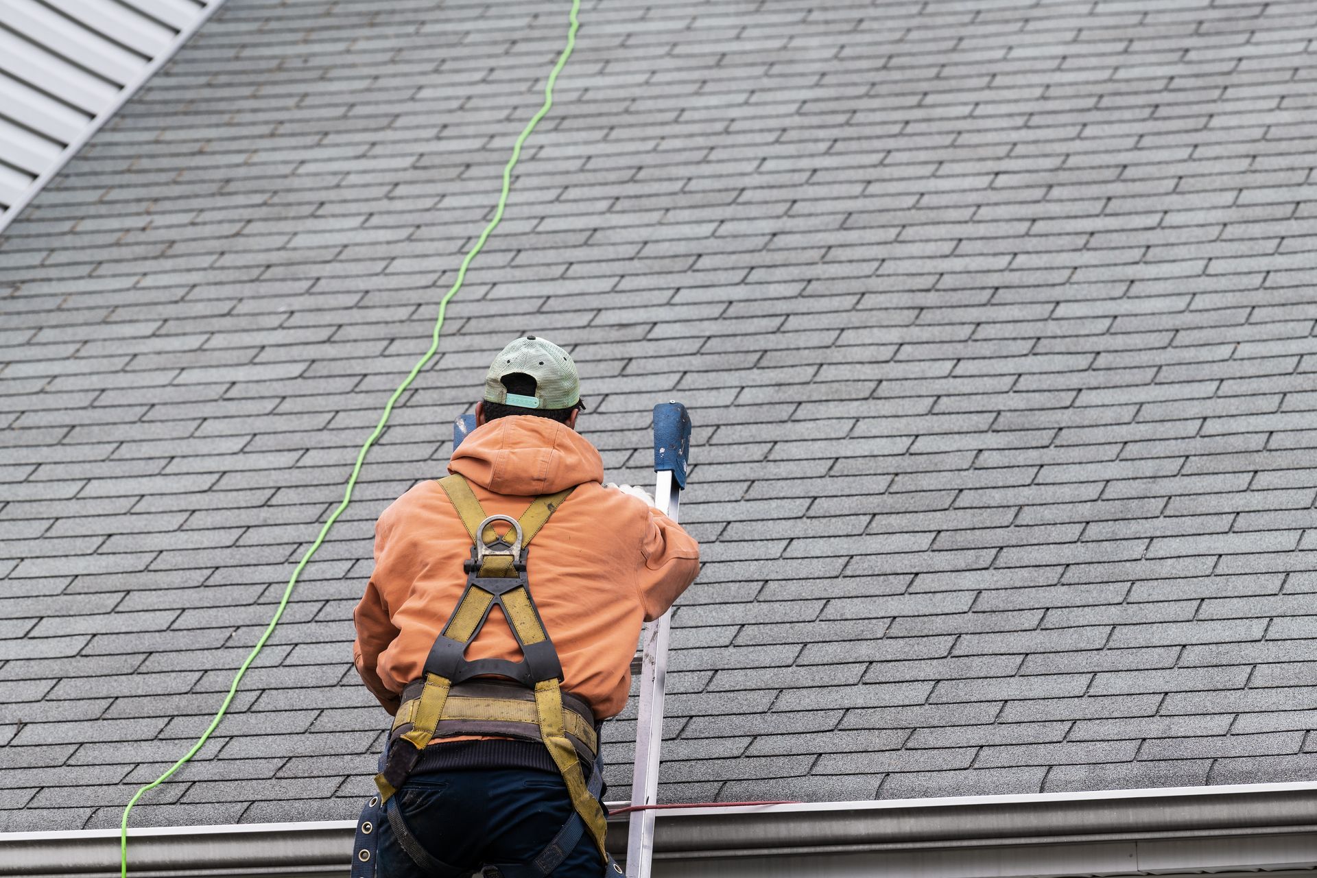 Worker in orange jacket and harness on a roof ladder, inspecting gray shingled roof.