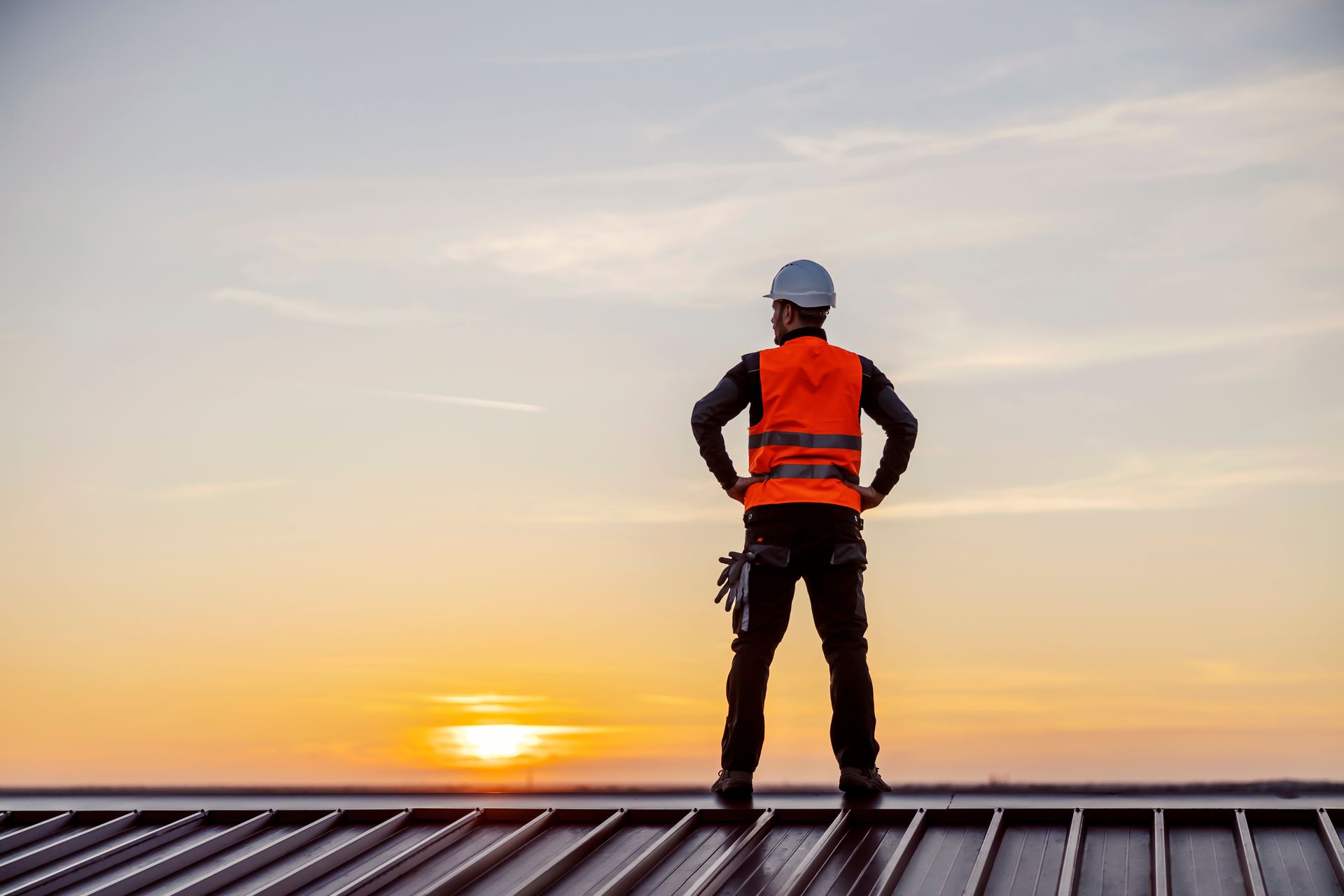 Roofing contractor in safety vest and hard hat standing on a metal roof at sunset.