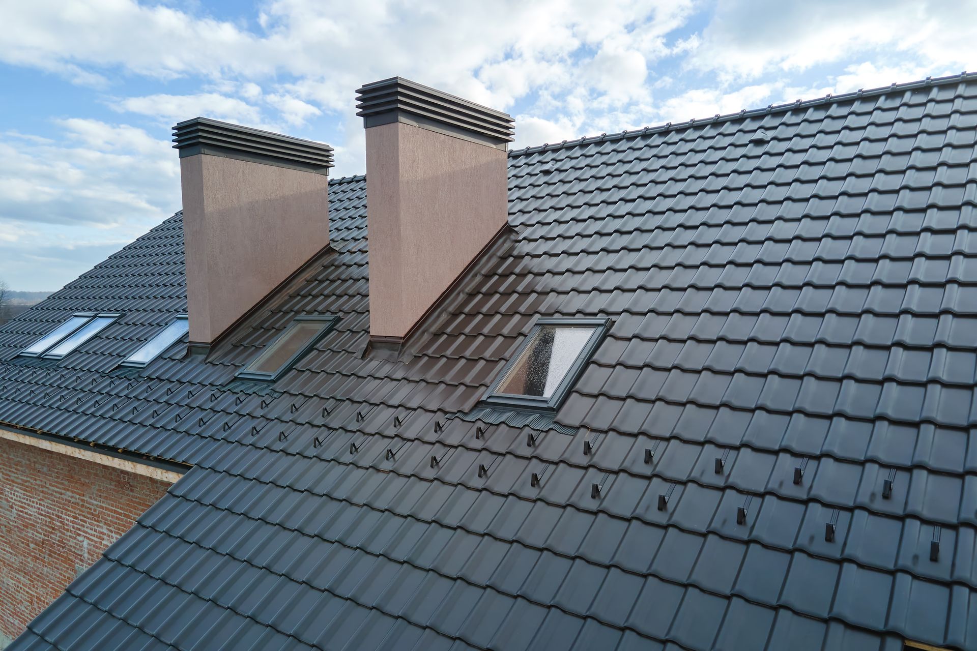 Dark gray tiled roof with two brick chimneys and skylights under a partly cloudy sky