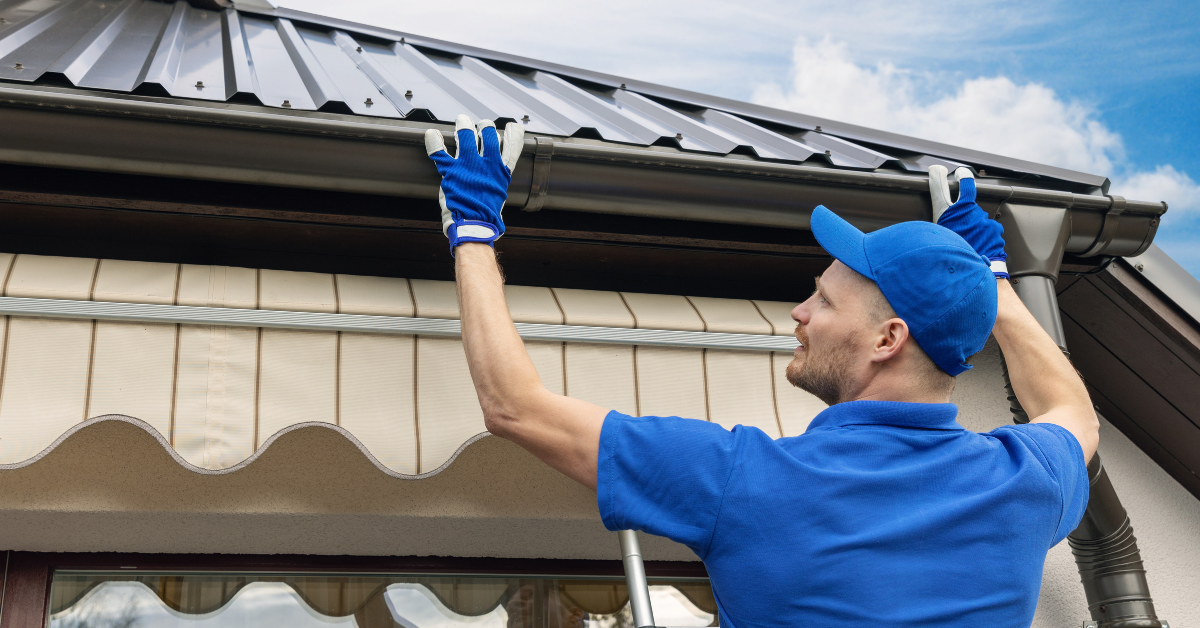 Worker in blue gloves and cap installing a metal roof gutter on a house