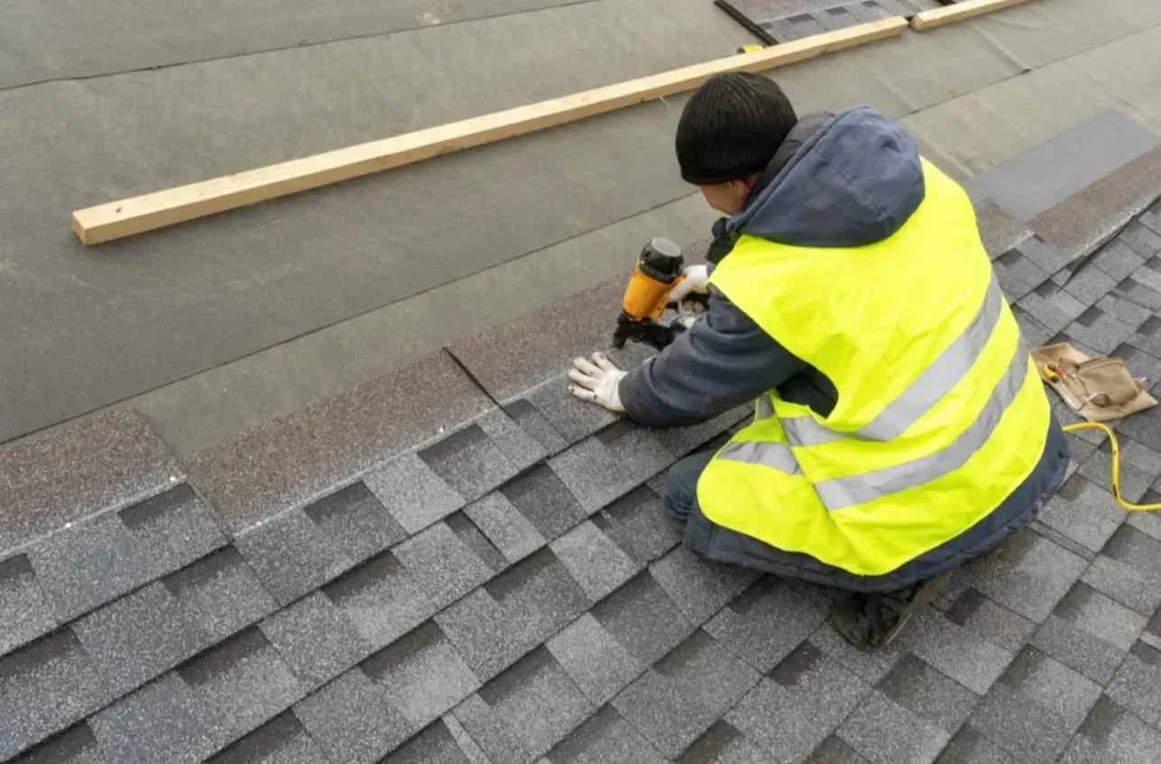 Worker installing shingles on a roof, wearing a yellow safety vest and using a nail gun