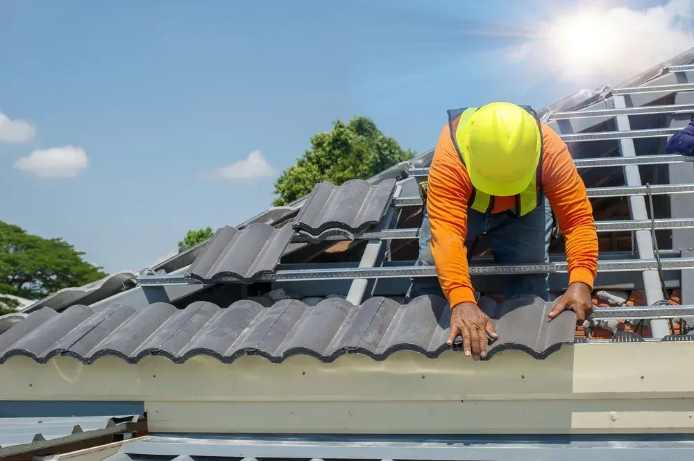 Worker in orange safety gear repairing a roof with gray shingles under a bright sky