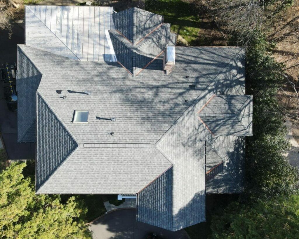 Aerial view of a gray-shingled house roof with skylights and chimney, surrounded by trees.