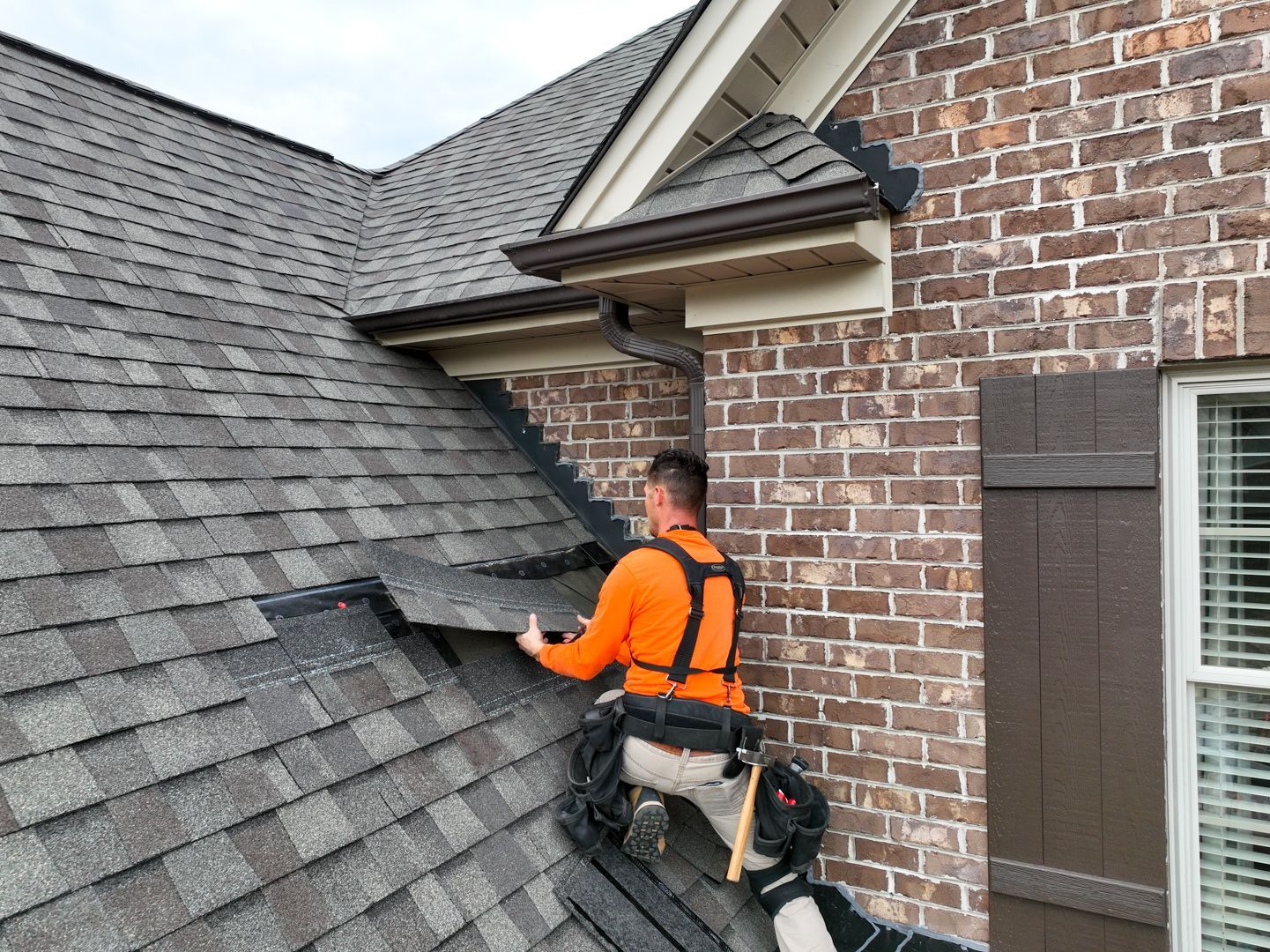 Worker in orange harness repairing gray shingles on a brick house roof