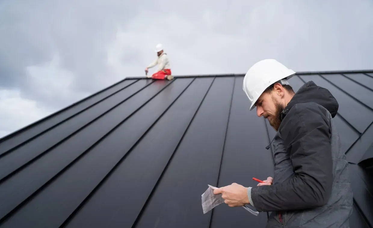 Worker in white hard hat inspecting a dark metal roof with another worker kneeling in the background
