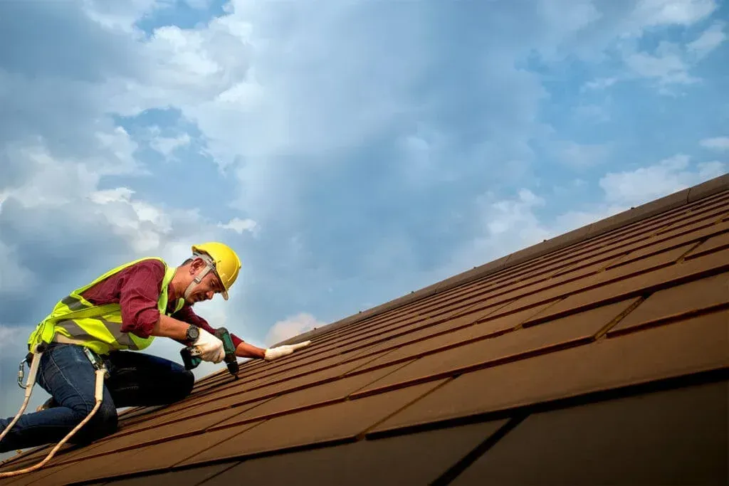 Worker in a yellow hard hat kneels on a rooftop, using a tool to repair brown shingles under a cloudy sky