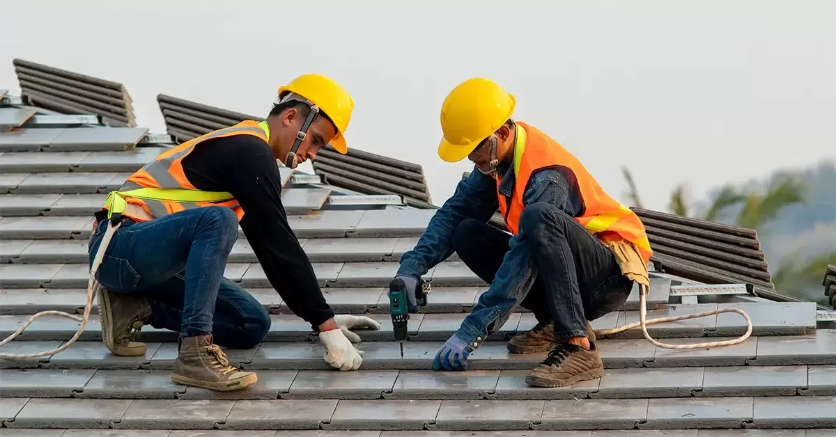 Two workers in yellow hard hats repairing a shingled roof with tools and safety vests