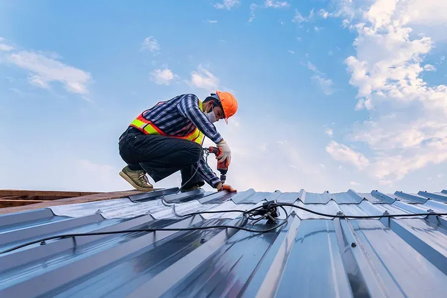 Roofer installing dark gray shingles on a sloped roof beside a brick chimney, with tools nearby