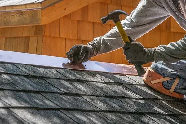 Roofer installing dark gray shingles on a sloped roof beside a brick chimney, with tools nearby
