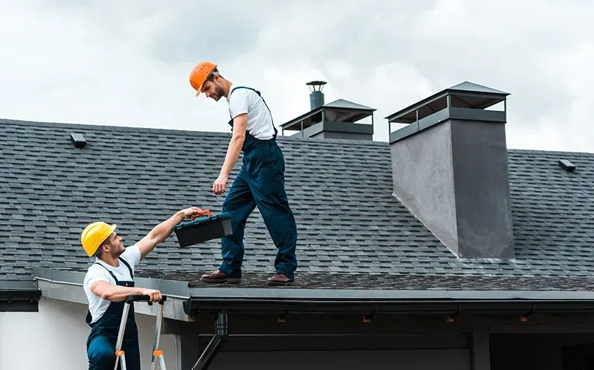 Two roofers in hard hats work on a shingled roof, one reaching down to help the other climb up.