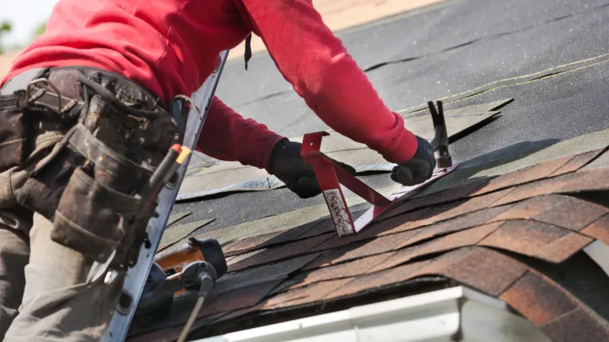 Worker in red hoodie repairing shingles on a rooftop with tools and safety harness