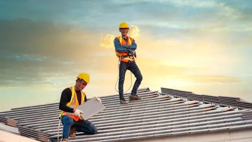 Two construction workers in hard hats inspect and repair a rooftop at sunset.