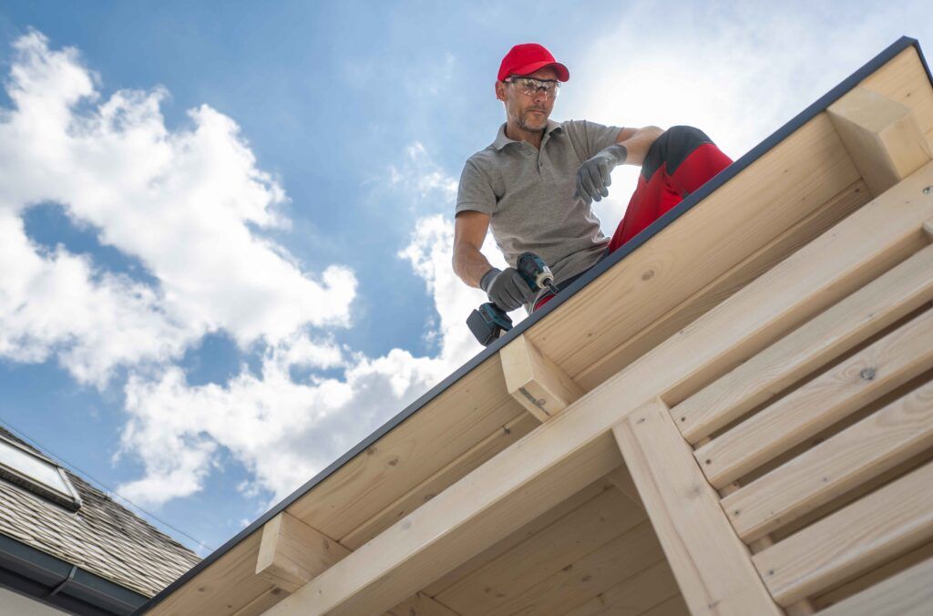 Roofer installing dark gray shingles on a sloped roof beside a brick chimney, with tools nearby