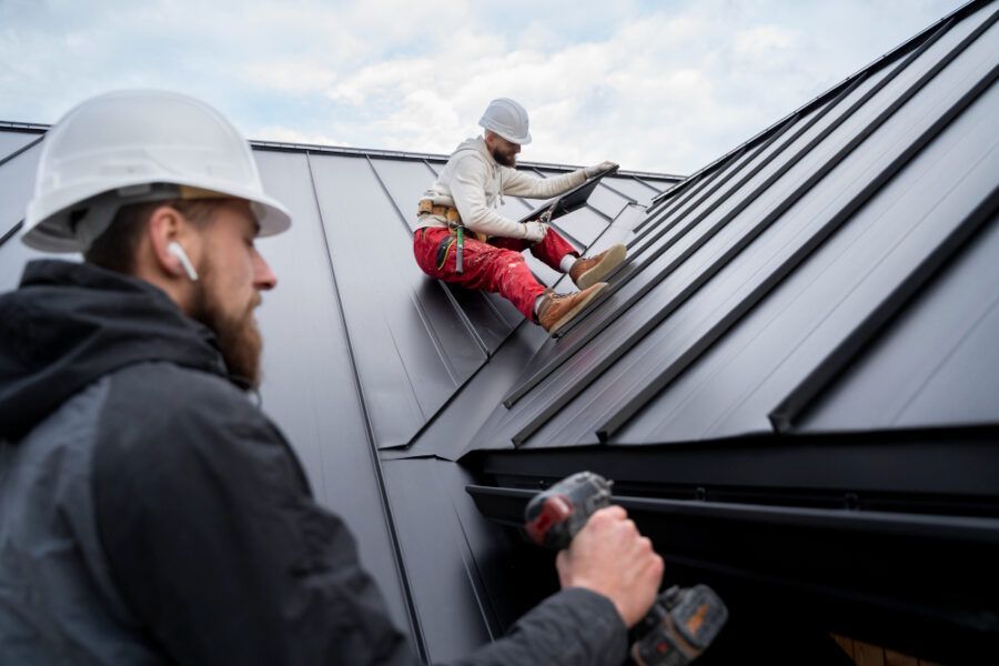 Two workers installing black metal roofing on a house, one on the roof and one below assisting