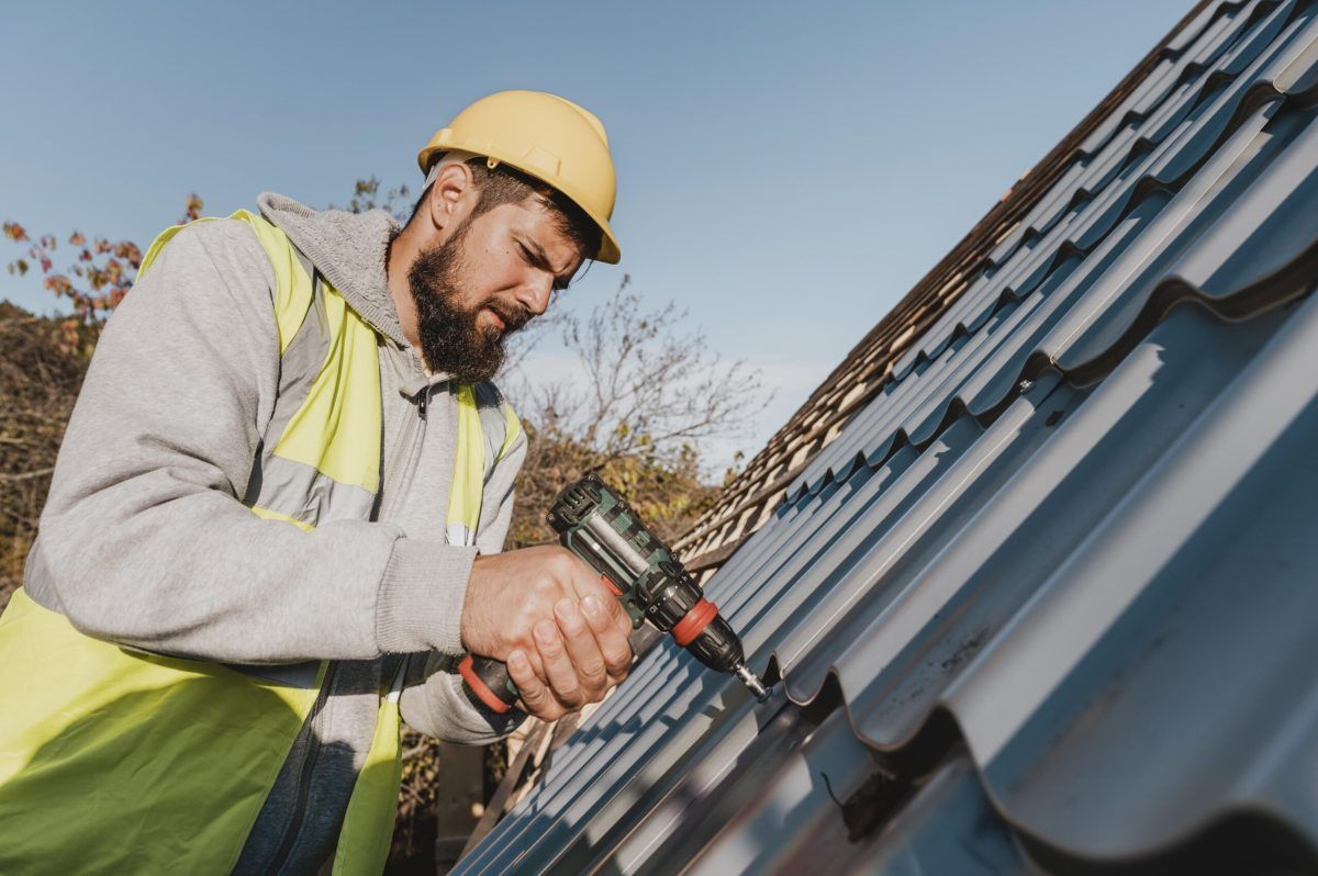 Worker in a yellow hard hat using a drill on a metal roof.