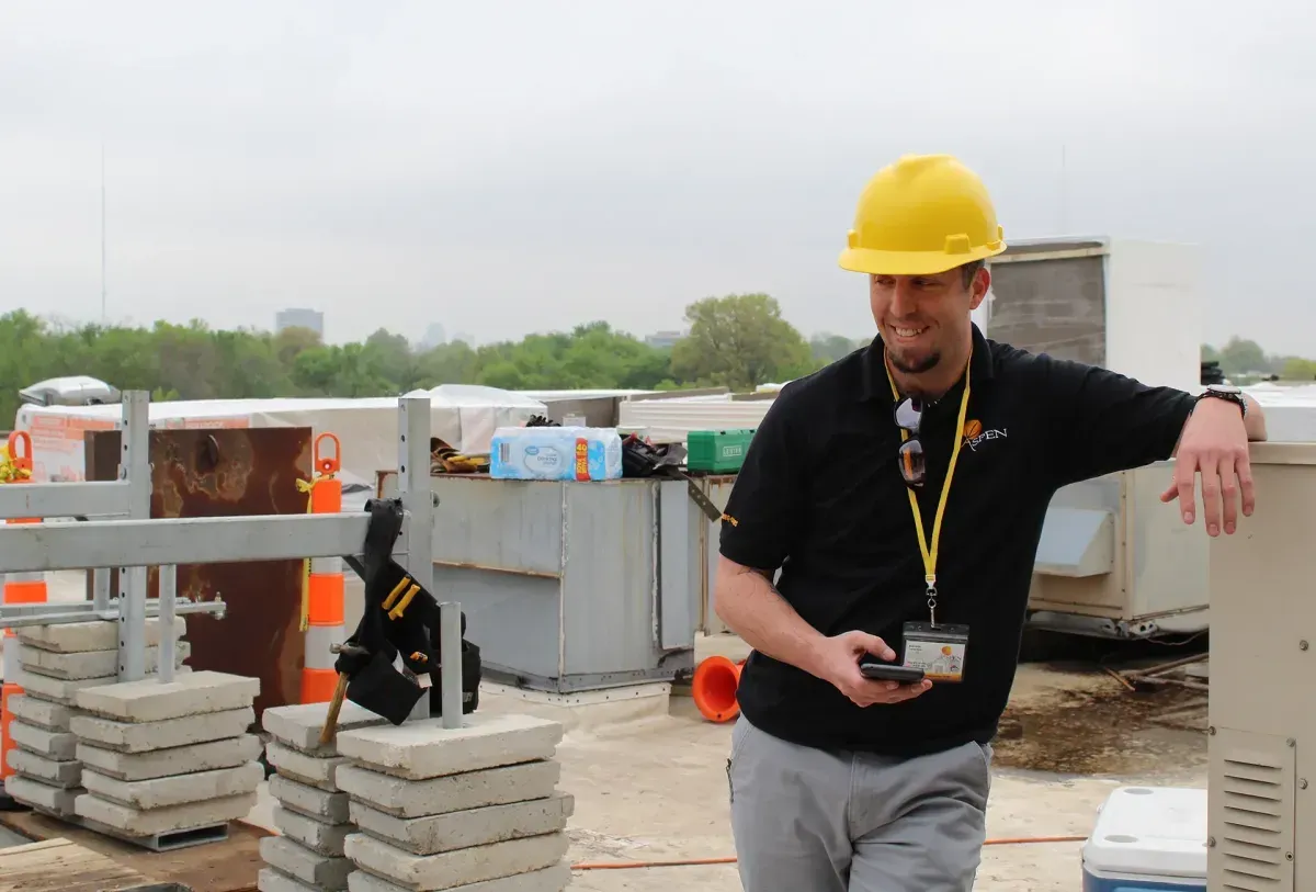 Construction worker in yellow hard hat leaning on a railing at an outdoor worksite, holding a phone.