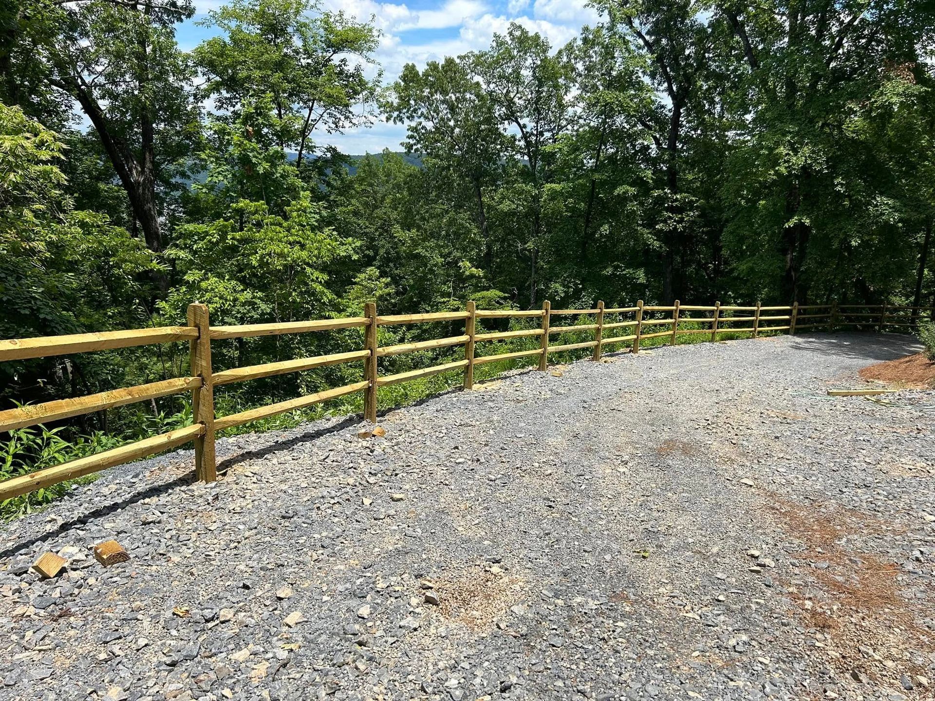 A gravel driveway curves behind a three-rail wooden fence in a wooded area.