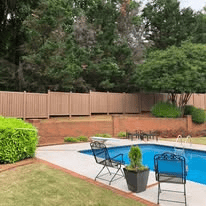 A backyard with a pool, fence, and greenery. Two chairs near the pool, and trees in the background.