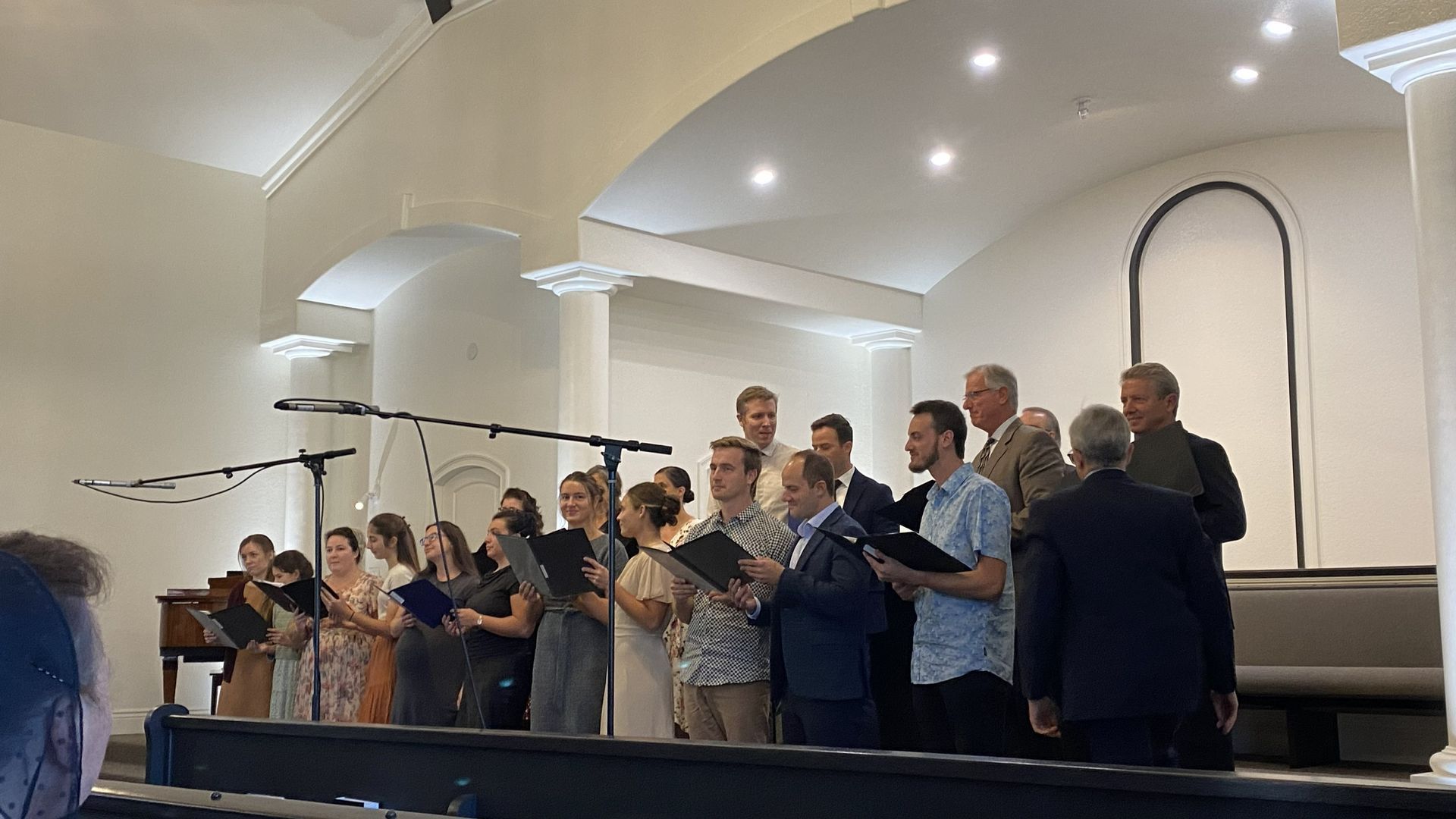 Choir singing on a church stage under bright lights, holding music folders at microphones