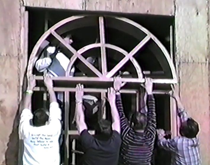 Several people lift a metal window grille during construction.