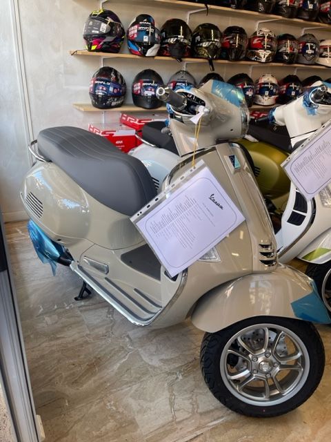 Scooter Vespa marrone con finiture cromate, parcheggiato all'interno di un negozio con caschi su uno scaffale sullo sfondo.