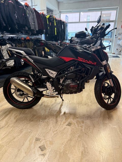 Motocicletta SWM nera e rossa in uno showroom con equipaggiamento motociclistico sullo sfondo.