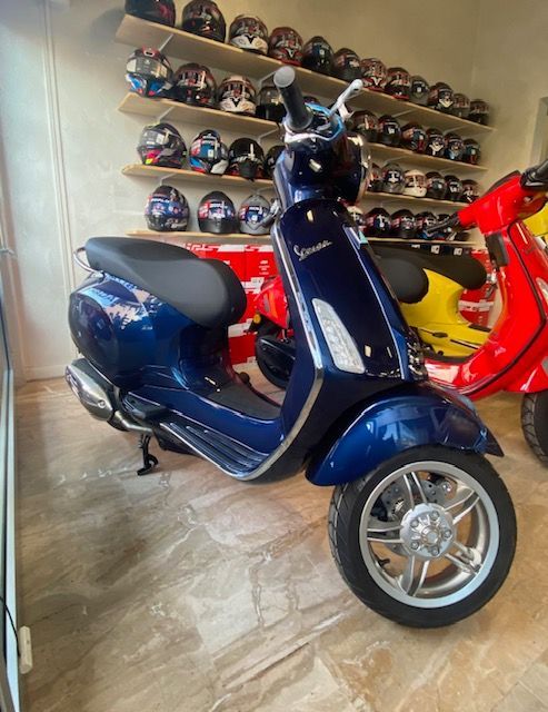 Uno scooter Vespa blu in uno showroom, finiture argentate, caschi sugli scaffali sullo sfondo.