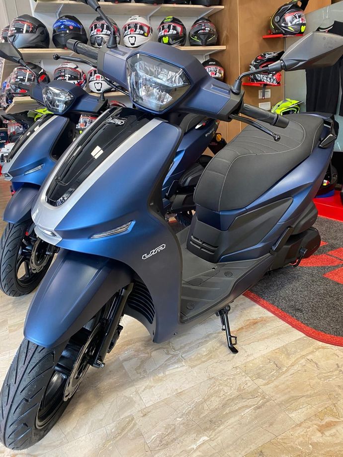 Monopattino blu e grigio esposto in un negozio, ruote nere, casco esposto sullo sfondo.