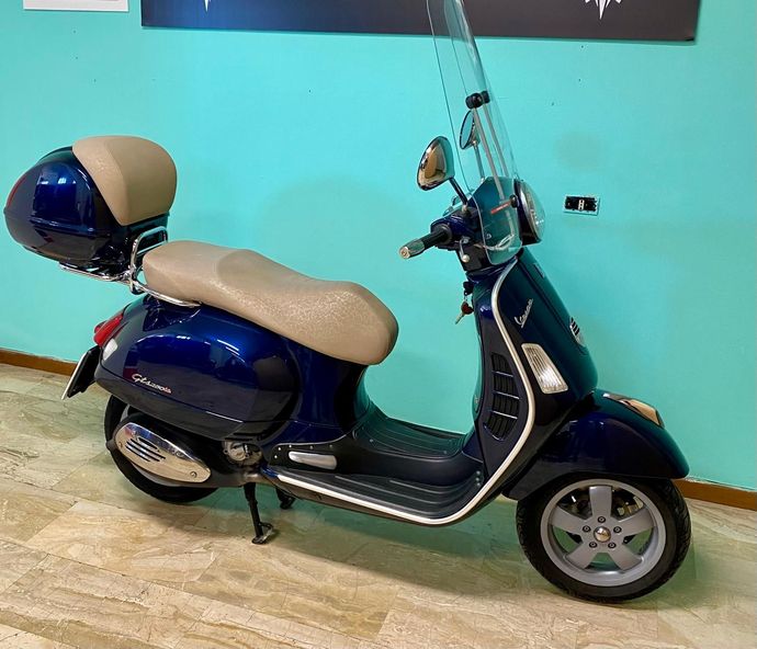 Scooter Vespa blu con sella e bauletto color cuoio parcheggiato su un pavimento piastrellato.