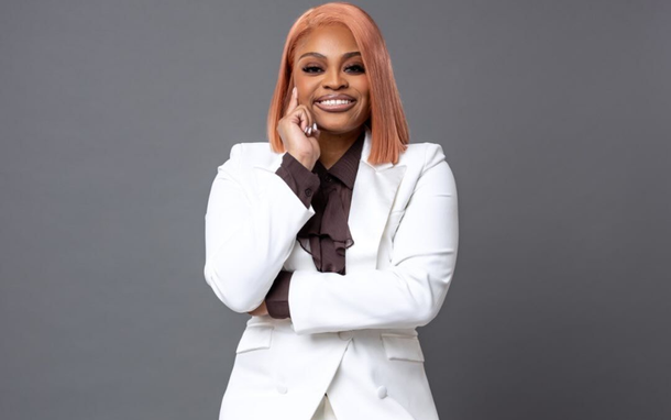 Woman in white blazer, brown blouse, smiling, pink hair, hand on face.
