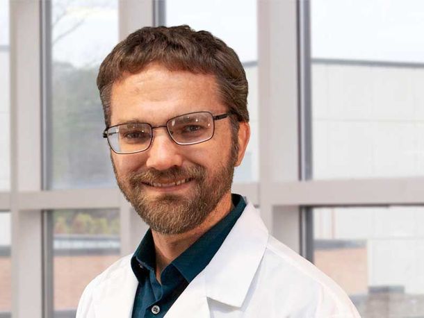 Smiling person in a white lab coat and glasses in an office or clinic setting