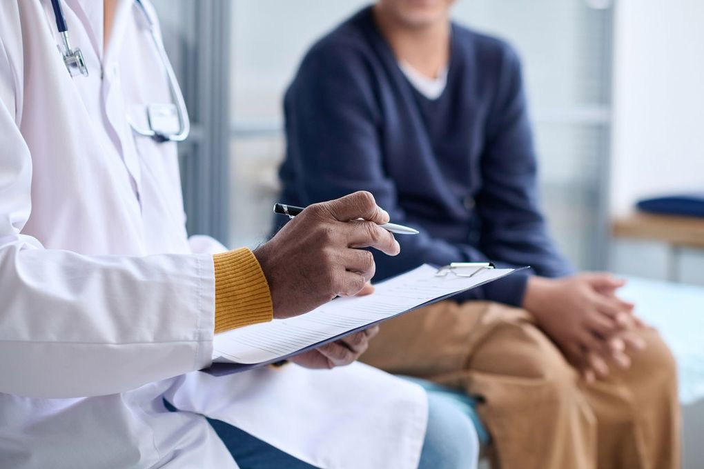 Doctor writing notes while speaking with a seated patient in a clinic office.