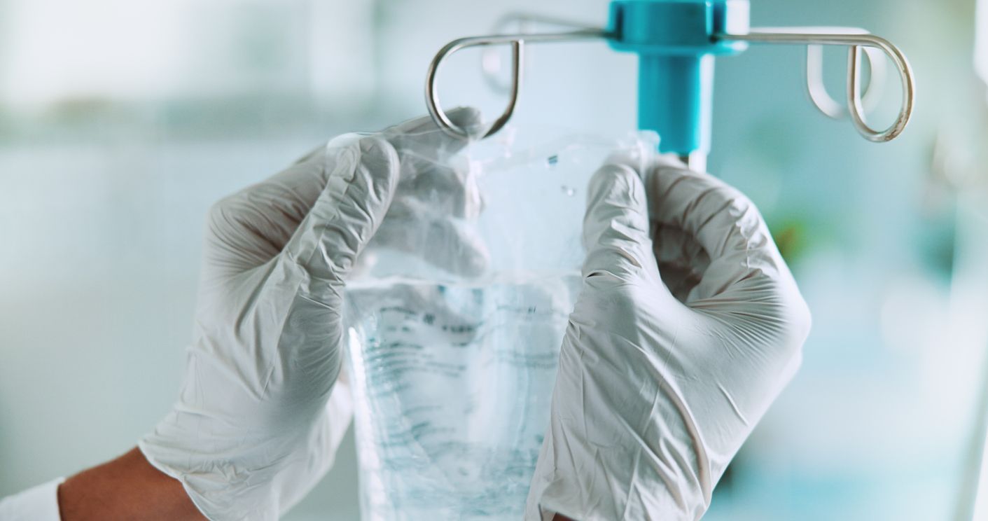 Gloved hands hanging a clear IV bag from a metal stand in a clinical setting