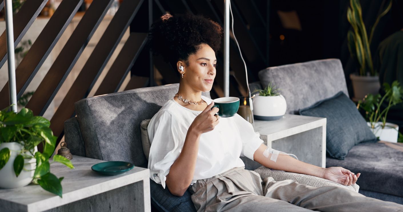 Person relaxing on a couch, holding a cup in a cozy living room with plants and soft pillows.