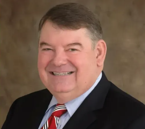 Smiling man in a dark suit, light blue shirt, and red striped tie against a brown background
