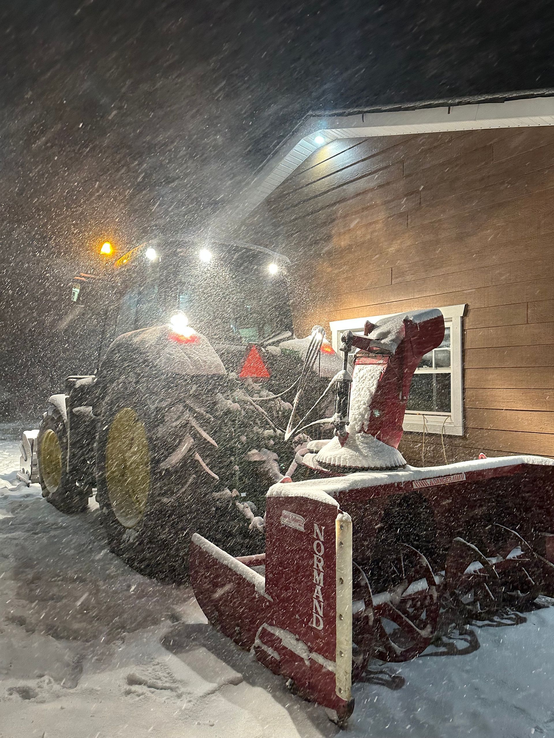 Un tracteur équipé d'une souffleuse à neige dégage la neige près d'un bâtiment, de nuit, éclairé par des projecteurs. Il neige abondamment.
