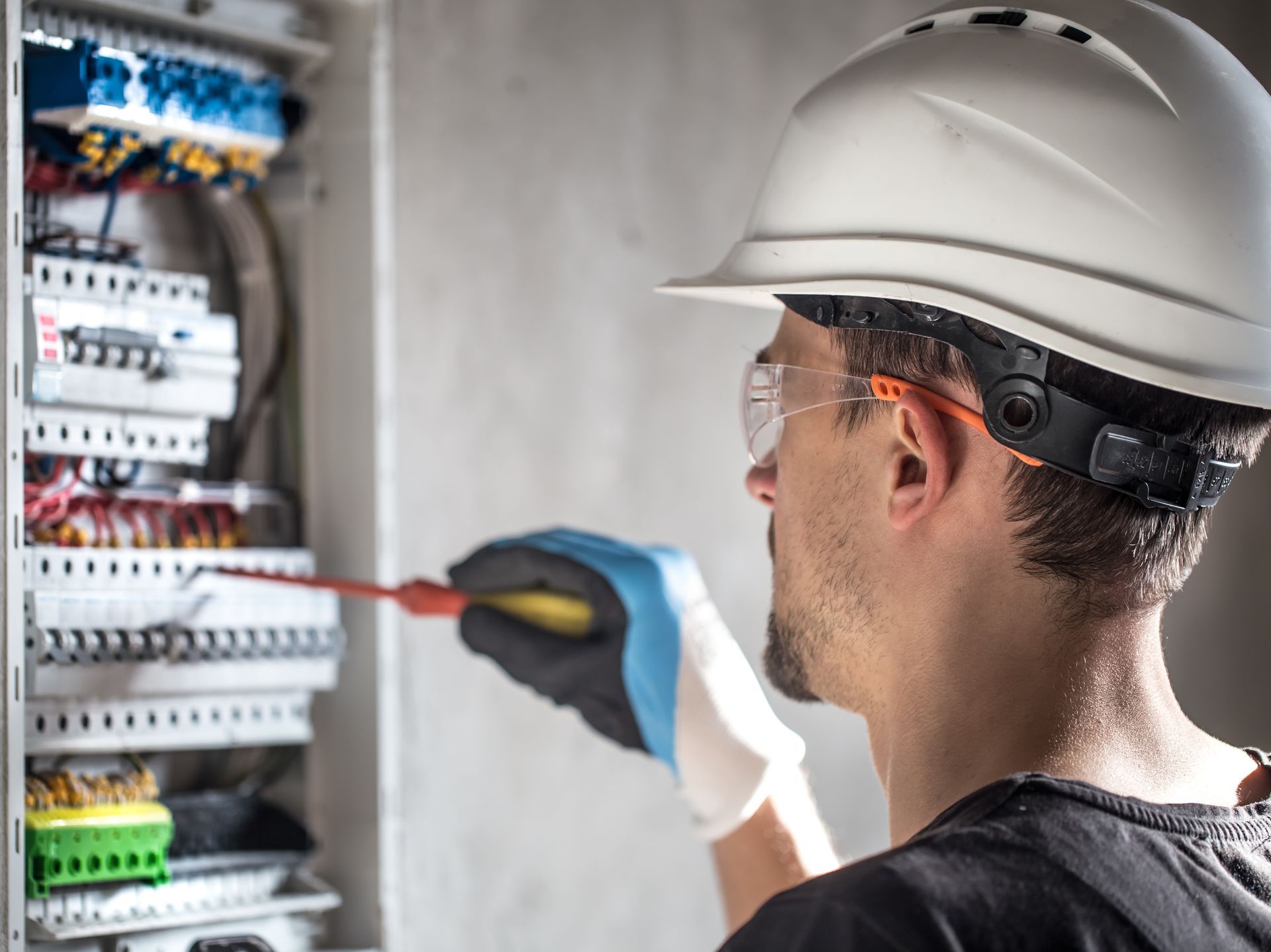 An electrician in a hard hat, safety glasses, and gloves is using a screwdriver on a circuit breaker panel.