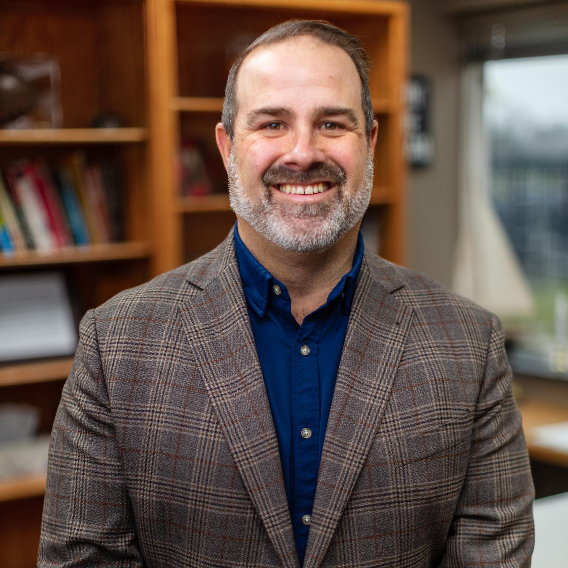 Smiling man in a plaid blazer and blue shirt standing in a book-lined office