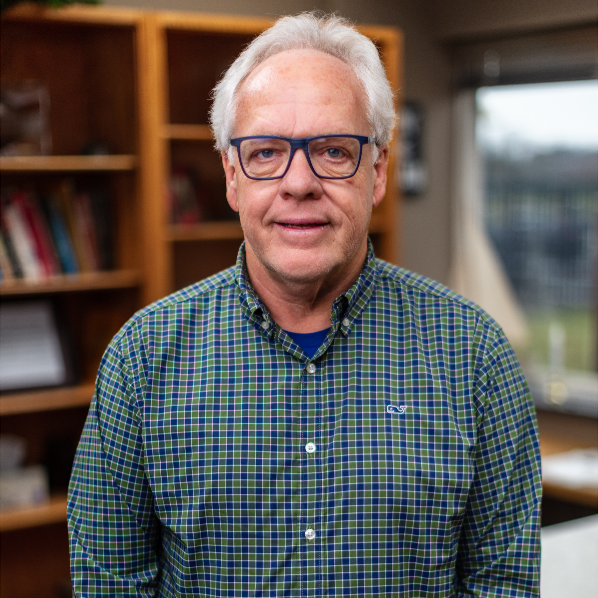Man wearing glasses and a blue-green plaid shirt, standing indoors by shelves and a window.