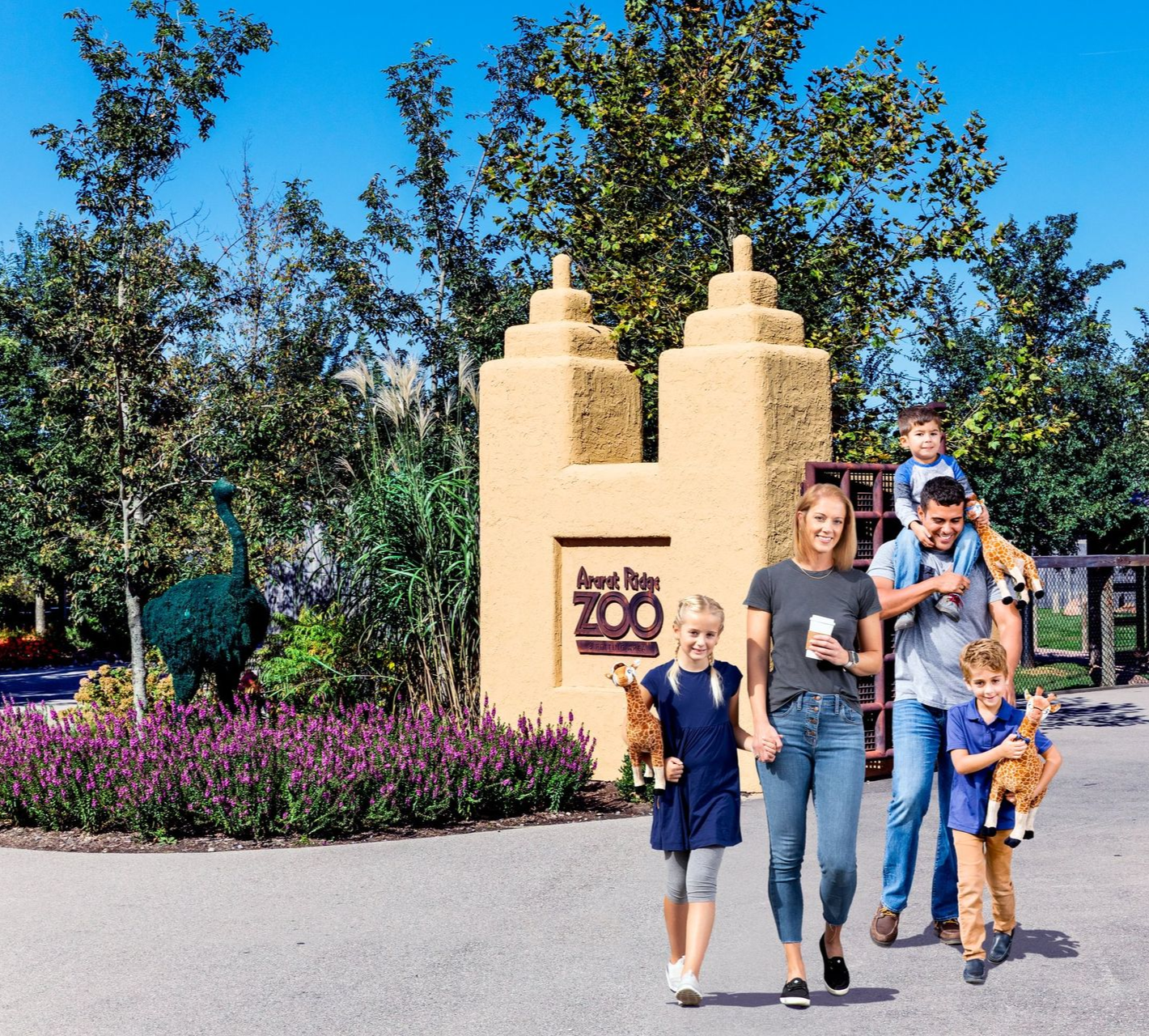 Family walking at a zoo entrance beside flower beds and a stone sign under a bright blue sky