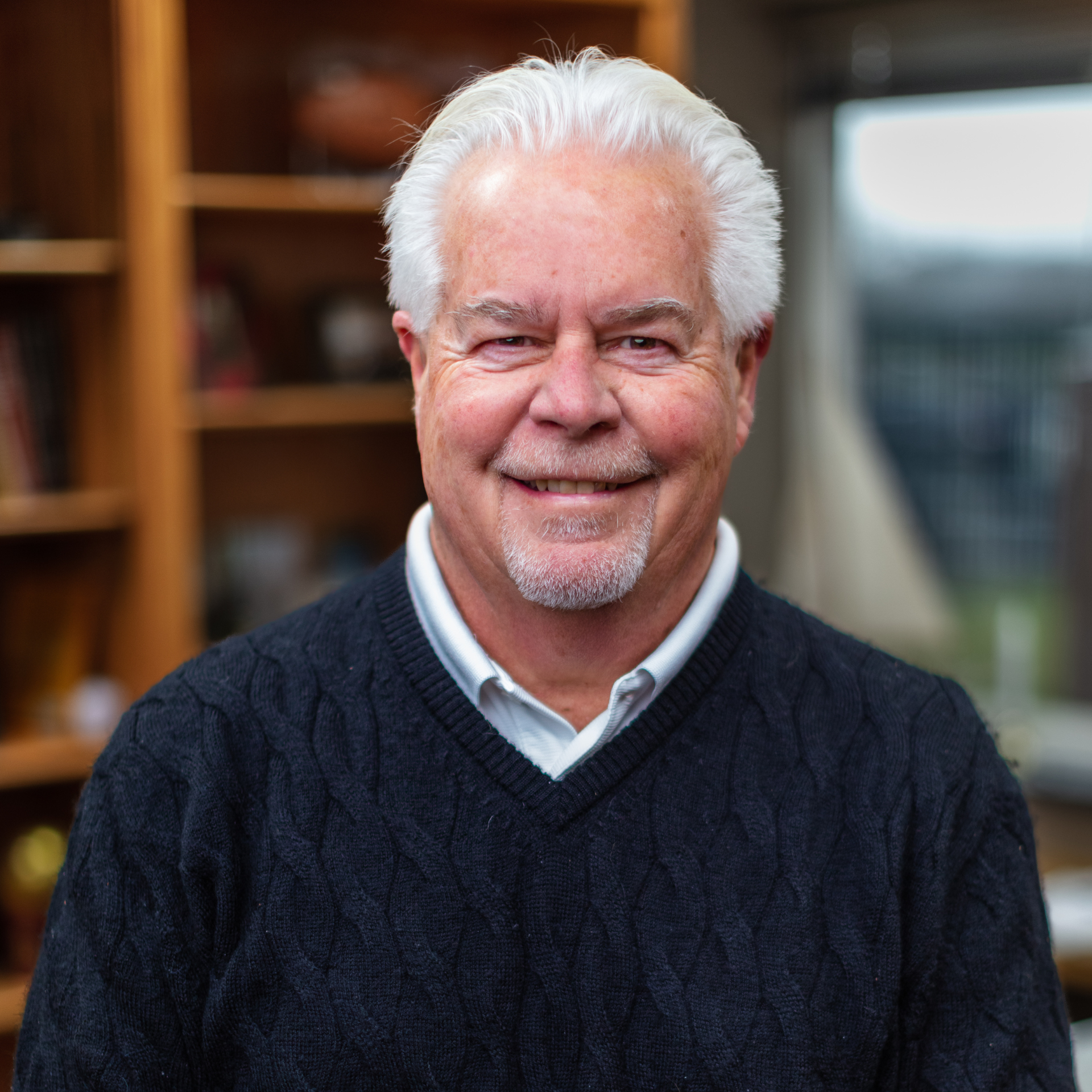 Smiling man in a navy sweater, seated indoors with a blurred wooden bookshelf background