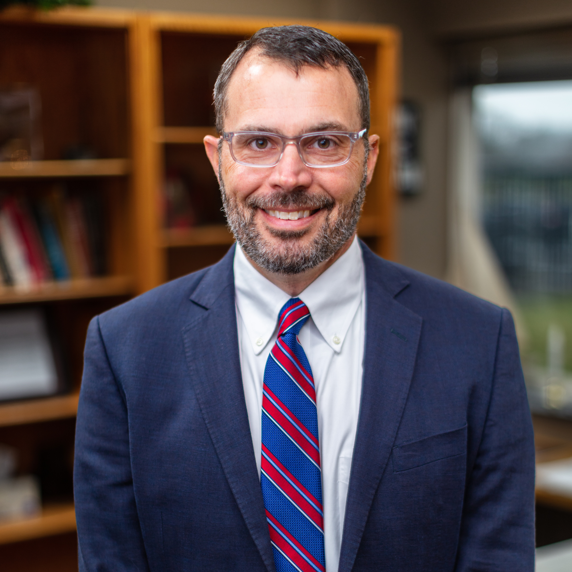 Smiling man in a navy suit, red striped tie, and glasses standing in front of bookshelves.