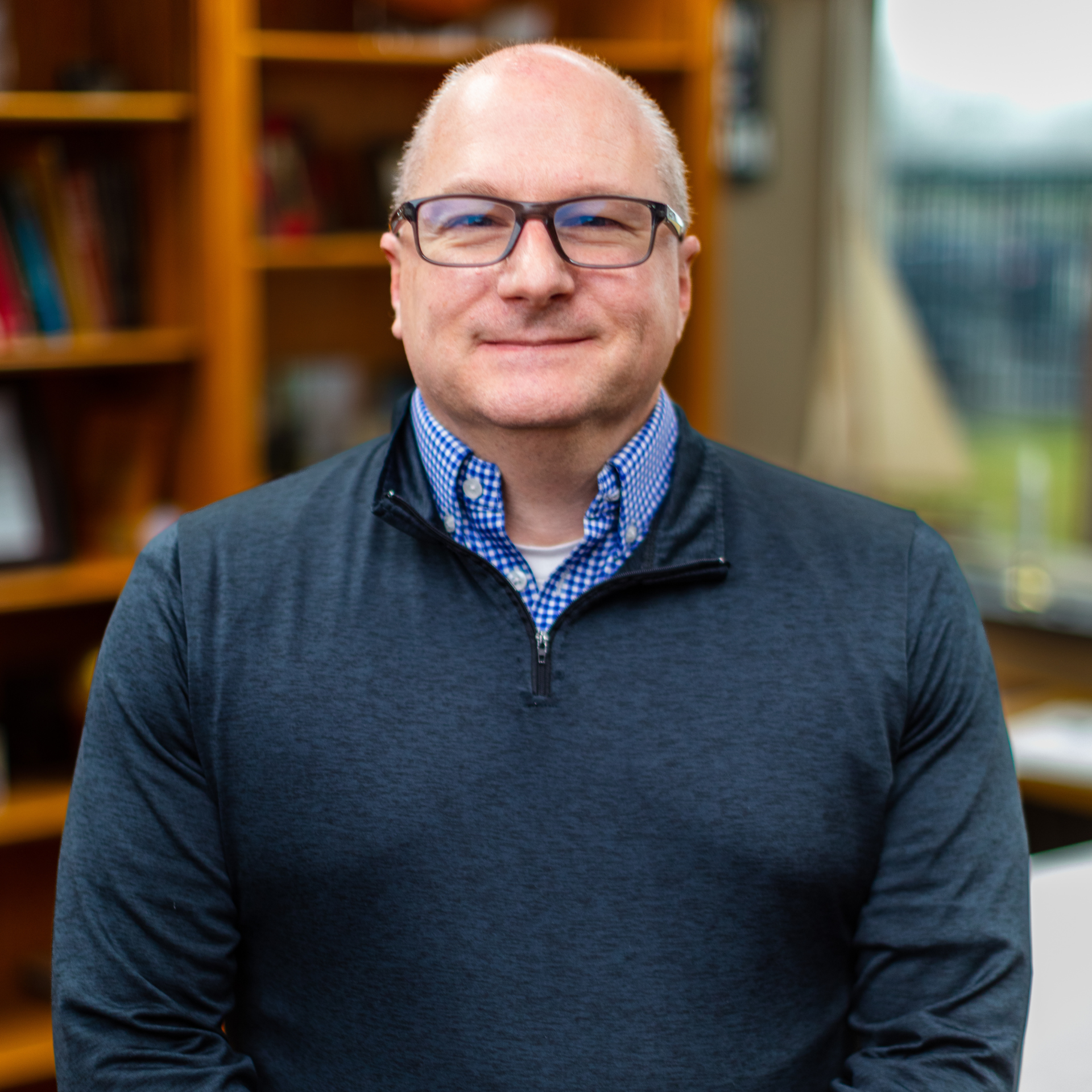 Portrait of a man in glasses wearing a dark sweater, standing indoors with bookshelves behind him