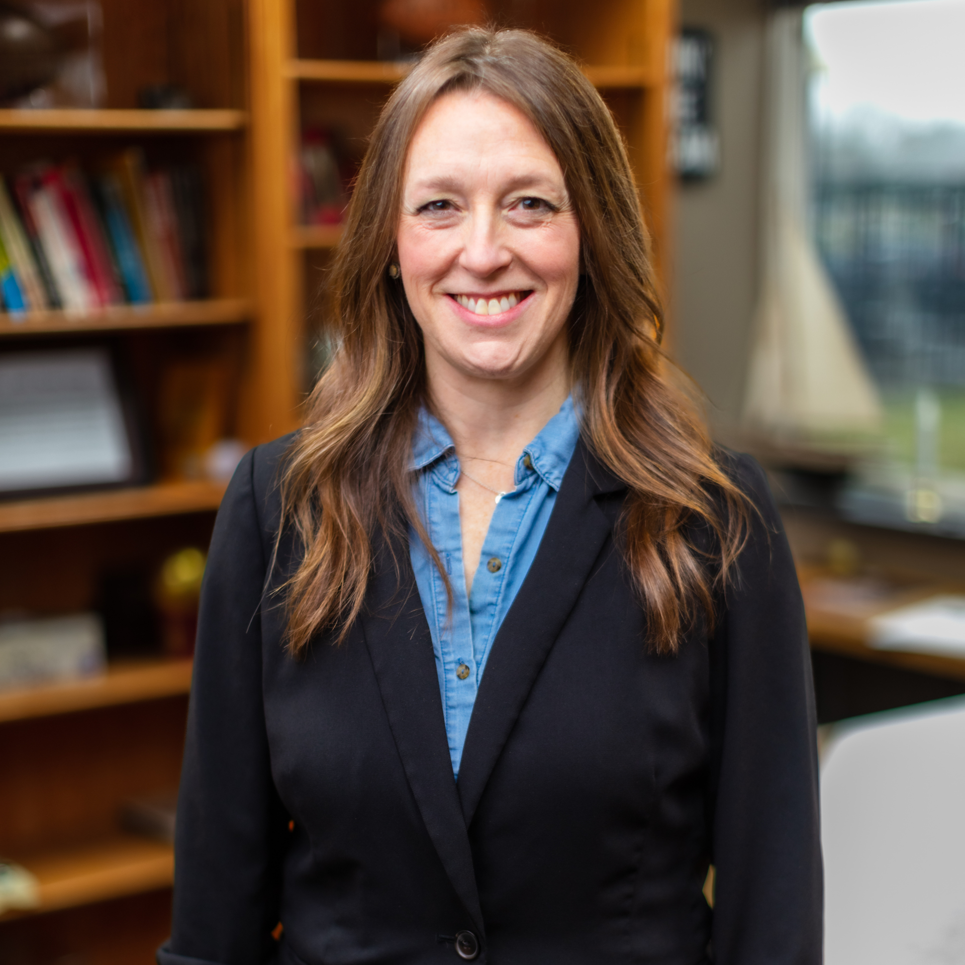Smiling woman in a black blazer and blue shirt standing in a library or office setting